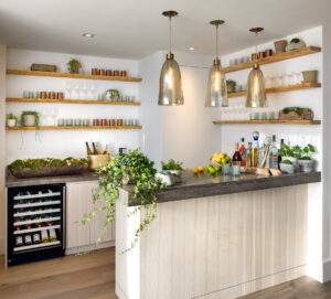 A modern bar with a dark counter, light wood base, open shelves, wine fridge, liquor, fruit, and plants.