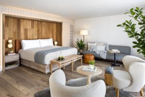 A contemporary bedroom with a wooden paneled headboard above a large bed, a gray sofa, and two cream armchairs on a wood floor.