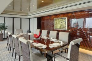 A formal dining area with a long polished wooden table and ten padded chairs. A high-gloss wood veneer wall features geometric patterns.