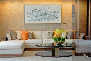 Light sectional sofa with orange and brown pillows, glass coffee table, and floral artwork on a textured gold wall.