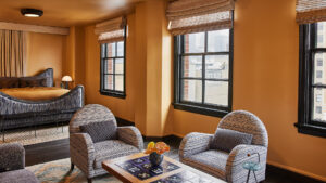 Ochre-walled suite with a sculpted queen bed, patterned area rug, two striped armchairs, a tile-inlay coffee table, and dark-framed city windows.