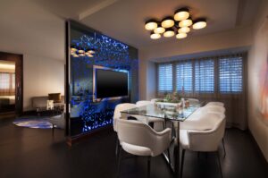 Modern dining area with glass table, white chairs, and a decorative partition wall with a TV, adjoining a living space.