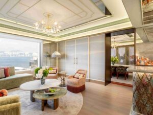 Living room with light green sofa and pink armchairs, nested stone coffee tables, glass chandelier, and window overlooking a harbor city.