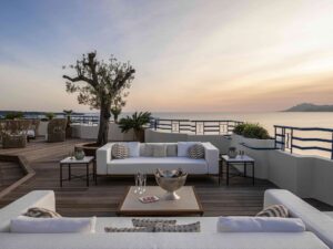 Outdoor terrace on a wooden deck with white seating, a large olive tree, and blue railings facing the ocean at sunset.