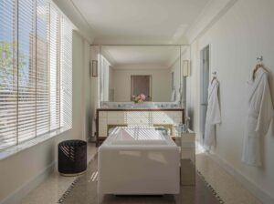 Bright bathroom with a central white soaking tub, double vanity under a wide mirror, and a window with horizontal slat blinds.