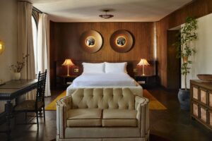 A bedroom with dark wood paneling behind a white-dressed bed, flanked by nightstands and lamps. A light tufted sofa is in the foreground.