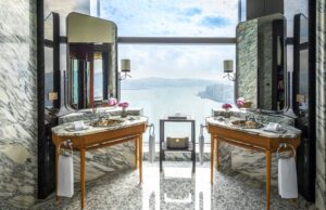 Double vanity bathroom with heavy veined marble walls and floors. Two wood vanities with metallic sinks flank a window overlooking a cityscape.