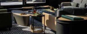 Close-up of a contemporary living area with gray and olive seating. Includes various tables, a tiered bar cart, and a rug with linear patterns near a dark-framed window.