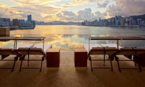Four sun loungers with white cushions line the edge of an infinity pool, which overlooks the Hong Kong skyline and harbor reflecting the golden sunset light.