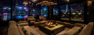 Nighttime view of a modern living room featuring a U-shaped fabric sofa, two armchairs, and a large coffee table with a central fire feature. Floor-to-ceiling windows reveal a harbor skyline.