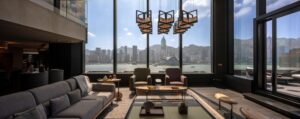 Modern duplex living room facing the Hong Kong skyline through full-height windows. Includes a gray sofa, fireplace coffee table, and terrace access.
