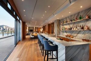 A luxurious open-plan bar, dining, and living area with a marble counter, blue bar stools, and a terrace overlooking a city.