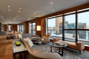 A modern living and dining room with floor-to-ceiling windows overlooking a city, harbor, and buildings. Grey sofa, wood floor.