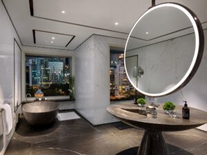 Modern bathroom with circular backlit mirror, unique pedestal sink, dark tub, and city skyline view at night.