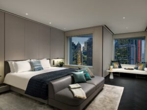 Modern hotel bedroom with large bed, gray sofa, and window seat. City skyline visible through large windows at night.