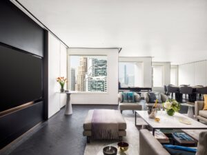 A modern living and dining room with dark wood floors, light walls, contemporary furniture, and large city-view windows.