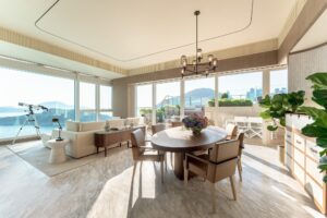 A bright living and dining room with large windows showing a bay and mountains, featuring a sofa, dining table, and outdoor terrace.