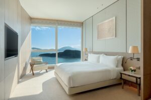 A modern bedroom with a large bed, armchair, and floor-to-ceiling windows overlooking the sea and mountains.