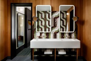 A modern bathroom with a double sink vanity, backlit mirrors, and a wavy-patterned tile backsplash against wood-paneled walls.