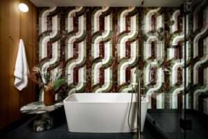 Modern bathroom with freestanding tub, glass shower, and a striking wall of green, brown, and white patterned marble mosaic.