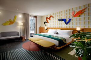 A vibrant bedroom with a king-size bed, abstract wall art, geometric wallpaper, a gray sofa, and a large plant.