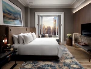 Bedroom with king bed, brown headboard, abstract art, textured walls, wood paneling with TV, patterned rug, and a window framing a skyscraper view.