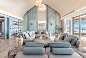 Indoor living room with light gray sectional seating, an arched ceiling, natural wood side tables, and ocean views through glass sliding doors.