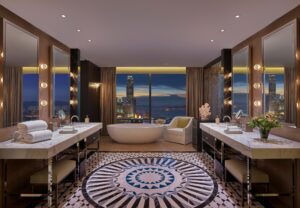 Bathroom with large window overlooking city and harbor, freestanding tub, two vanity sinks, and patterned floor.