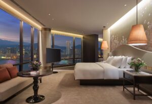 A luxury bedroom with a large bed, sofa, and TV, offering a panoramic view of a city skyline and harbor at night.