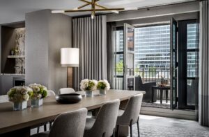 Modern dining room with long table, upholstered chairs, chandelier, open balcony, and city buildings outside.