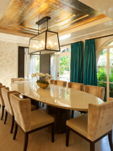 Formal dining room with a glossy light oval table set for twelve, lit by a lantern chandelier. Arched glass doors open to a garden.