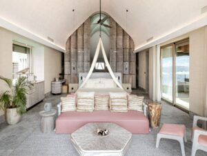 Interior of a bedroom with a king bed, pink daybed, and vaulted ceiling. Sliding glass doors open to an overwater deck and ocean view.