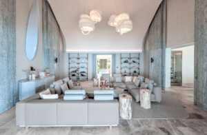 High-ceilinged lounge featuring a large gray modular sofa, natural wood stump side tables, and four braided rope chandeliers. Textured walls and marble floor.