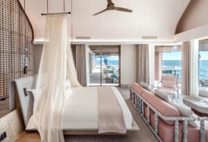 Light-toned bedroom with a king bed, sheer canopy, and vaulted ceiling. It has large glass doors opening to a deck overlooking the turquoise ocean.