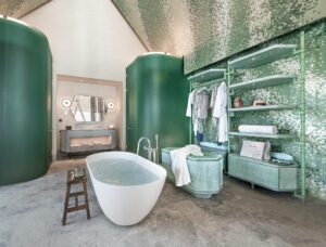 A spacious bathroom with a vaulted ceiling and green mosaic tiles. It includes a freestanding tub, double vanity, and open shelving for robes and clothes.