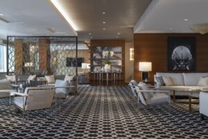 Large lounge area with light sofas, armchairs, and patterned carpet. Features wood walls and a metal partition.