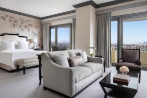 Bedroom featuring a king bed, floral accent wall, light gray sofa, and glossy dark wood coffee table facing balcony doors with a city view.