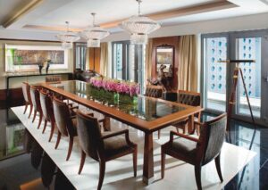 Formal dining room with long wooden table, ten chairs, chandeliers, floor-to-ceiling windows, and a standing telescope.