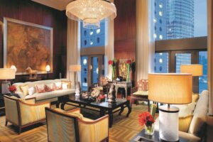 A luxury living room with a large chandelier, dark wood panels, a sofa, armchairs, and tall windows showing city buildings.