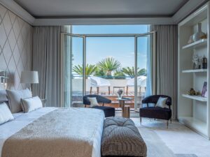 Bedroom with king-sized bed, quilted headboard, and built-in white shelves opening to a sunlit terrace with palm trees and outdoor seating.