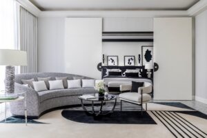 Monochrome sitting area with a curved gray sofa and black coffee table, defined by a graphic rug, opening to a bedroom via white sliding doors.