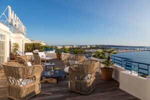 Rooftop deck with wooden flooring, woven chairs, and white sun loungers facing the Mediterranean Sea and a large marina.