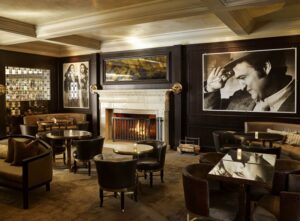 Upscale bar with dark paneling, a marble fireplace, leather seating, and small round tables. Large black and white portraits adorn the walls.