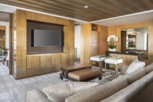 A modern living room with a large light sofa, a brown ottoman, and wood paneling, featuring a TV and a bar area.