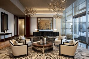 A luxurious lobby with light armchairs, a round coffee table, ornate chandeliers, a large reception desk, and glass doors.
