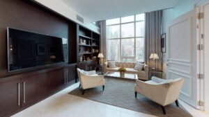 A luxurious living room with a large TV, dark wood shelving, a sofa, two chairs, and city views through large windows.