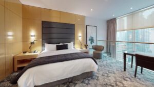 Modern bedroom with large bed, wood-paneled wall, nightstands, and armchair next to glass railing and city-view windows.