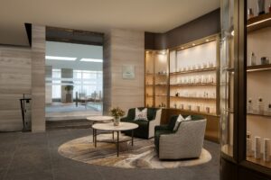 Spa waiting area with green armchairs, two coffee tables, illuminated product display cases, and an indoor pool visible through a glass wall.