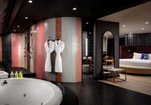 A modern bathroom with a white whirlpool tub and striped mosaic shower area, leading into a bedroom.