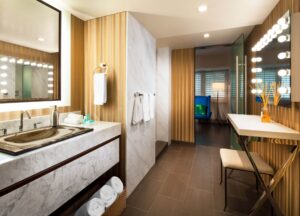 Modern bathroom with marble vanity, integrated sink, large mirror, ribbed gold walls, lighted makeup mirror, and tiled floor.